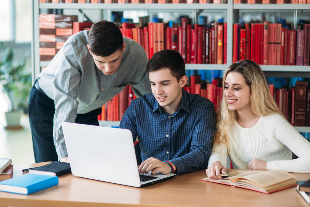 University students sitting together at table with books and laptop. Happy young people doing group study in library
